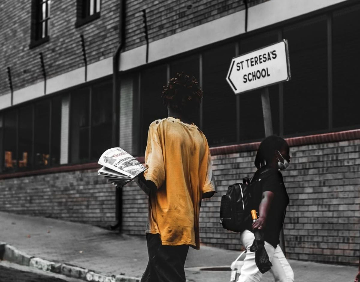 Newspaper seller walking past a school sign on a Johannesburg street