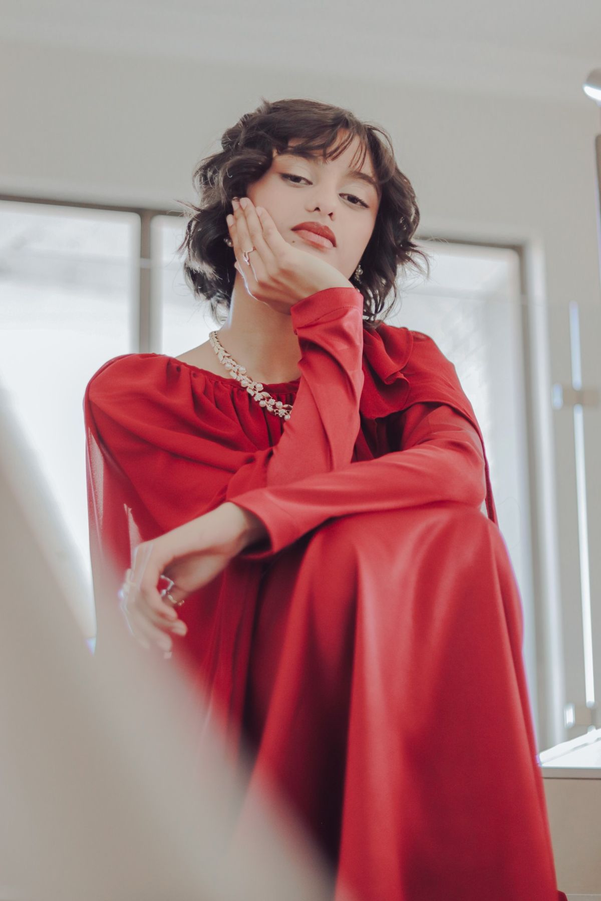 Model in a red dress seated by a window, resting her chin on her hand