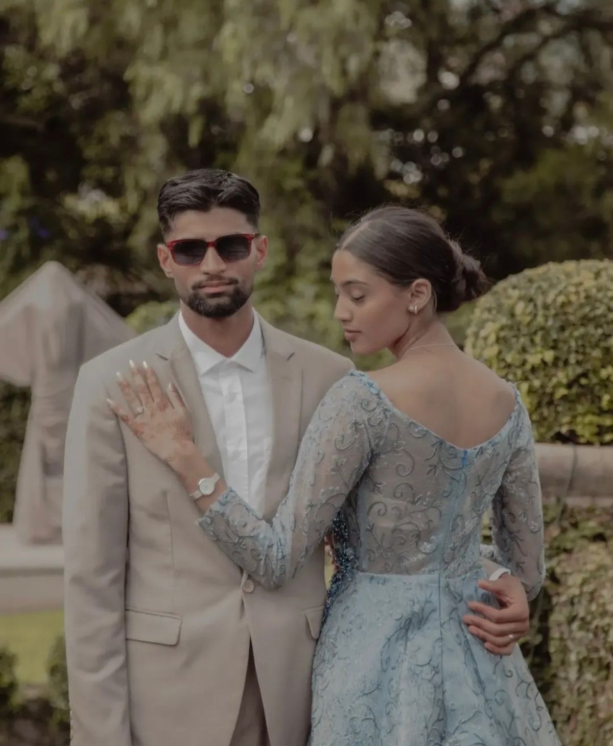 Couple in a garden, she in a blue lace gown with her hand on his shoulder