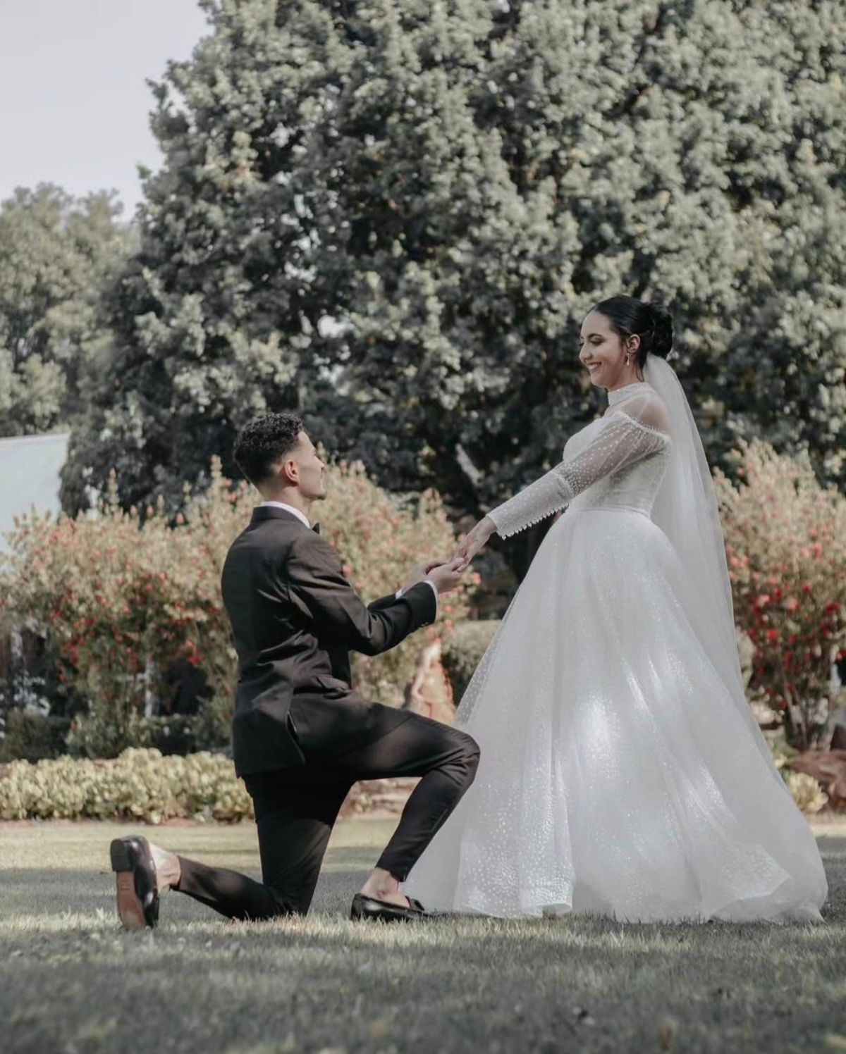 Groom kneeling on the lawn and holding the bride’s hand